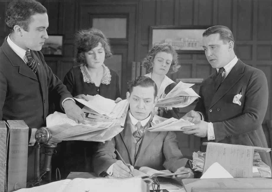 Film-Szene 30er Jahre: Angestellte mit verschiedenen Anliegen umringen einen Mann am Schreibtisch.